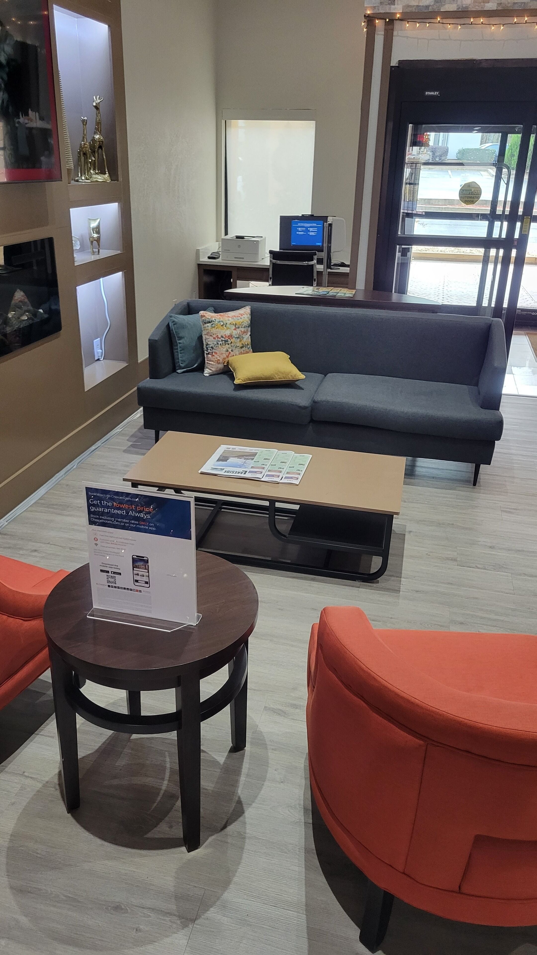 A modern lobby with a grey sofa, orange armchairs, a coffee table, and a desk with a computer.
