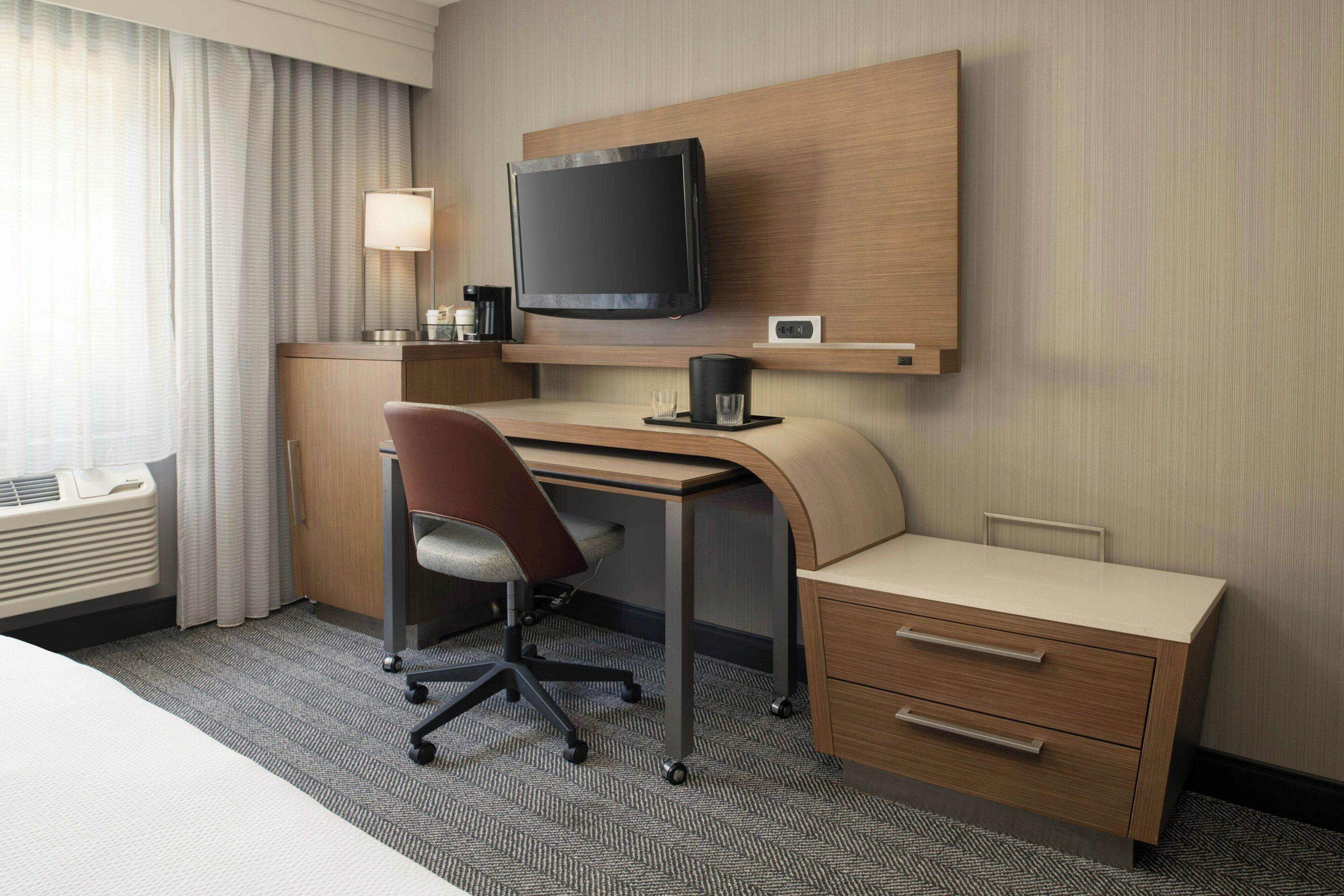 A hotel room with a bed, a desk with a chair, a television, a lamp, a coffee maker, and a small drawer unit.