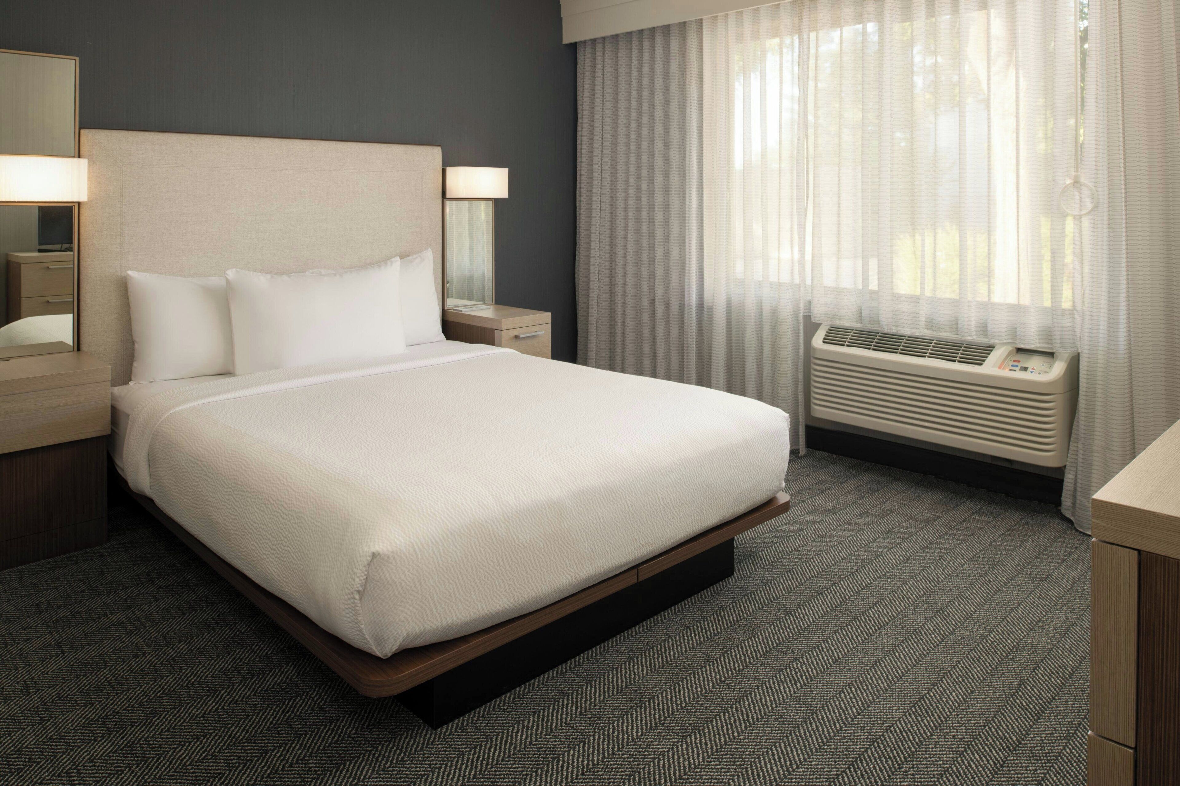 Habitación de hotel con una cama grande, una mesita de noche con lámpara, una ventana con cortinas y un aire acondicionado de pared.