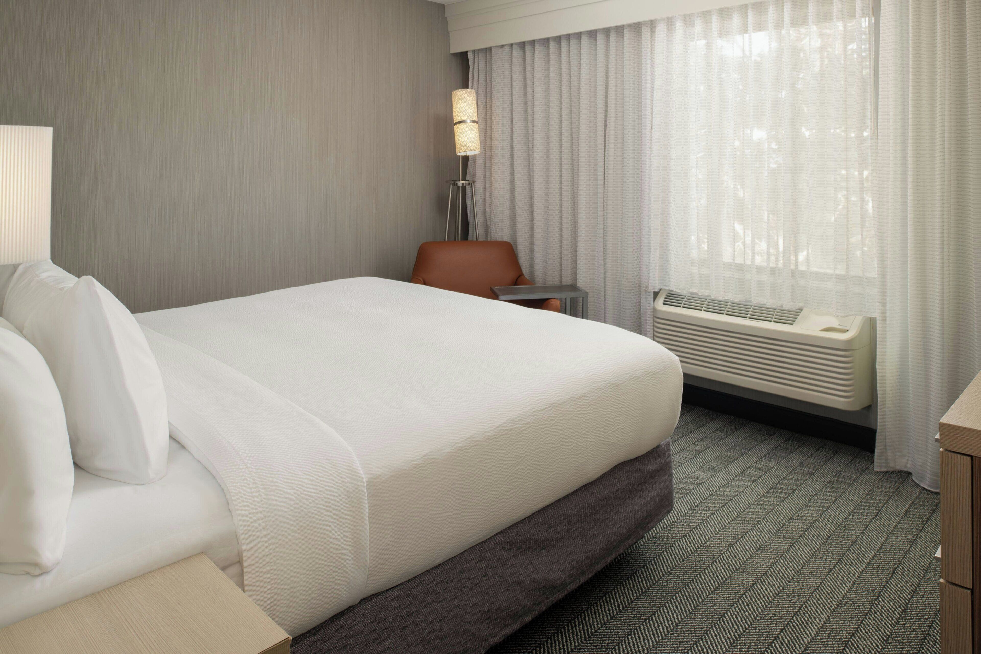 Habitación de hotel con una cama grande, un escritorio, una silla, una lámpara y una ventana con cortinas.