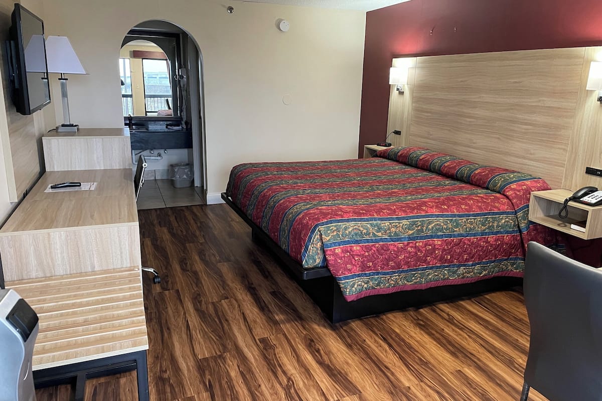 A hotel room with a bed, a desk, a chair, a TV, and a bathroom visible through an archway.