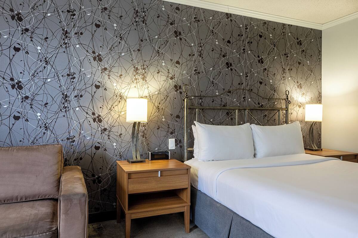 A hotel room with a bed, two bedside tables with lamps, a wooden nightstand, and a brown armchair.
