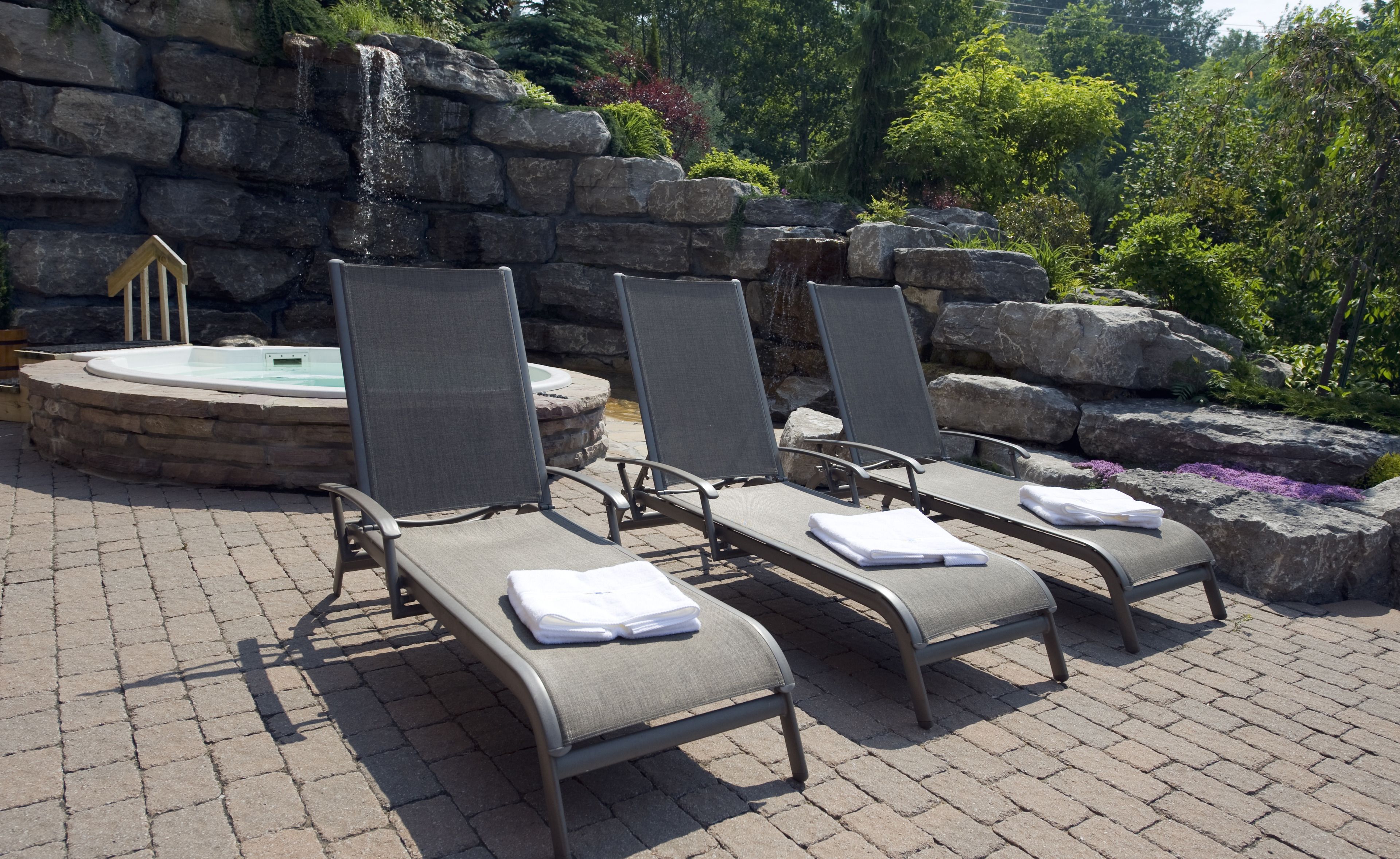 Three lounge chairs with towels, a hot tub, and a waterfall backdrop.