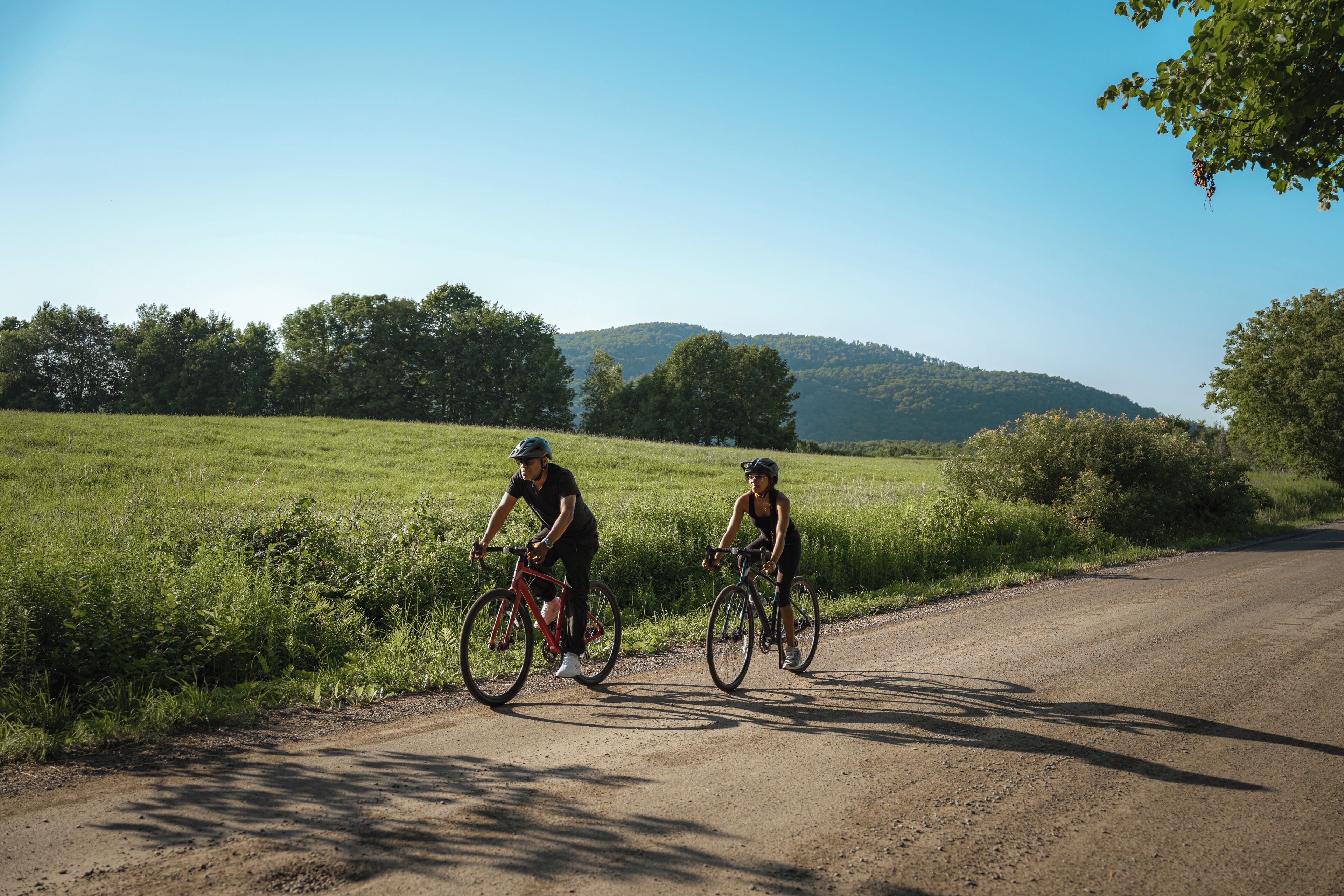 Dos ciclistas en una carretera rural con un campo verde y colinas al fondo.
