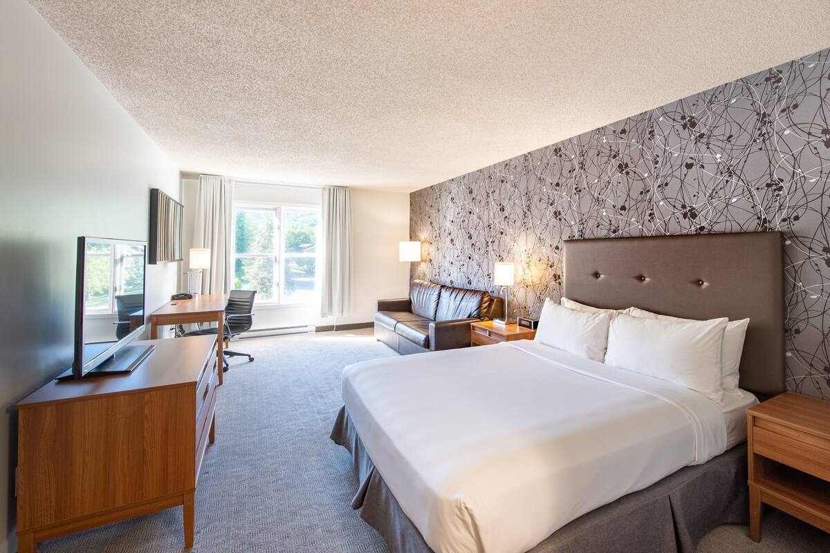 A hotel room with a large bed, a desk with a flat-screen TV, a sofa, and a window with curtains.