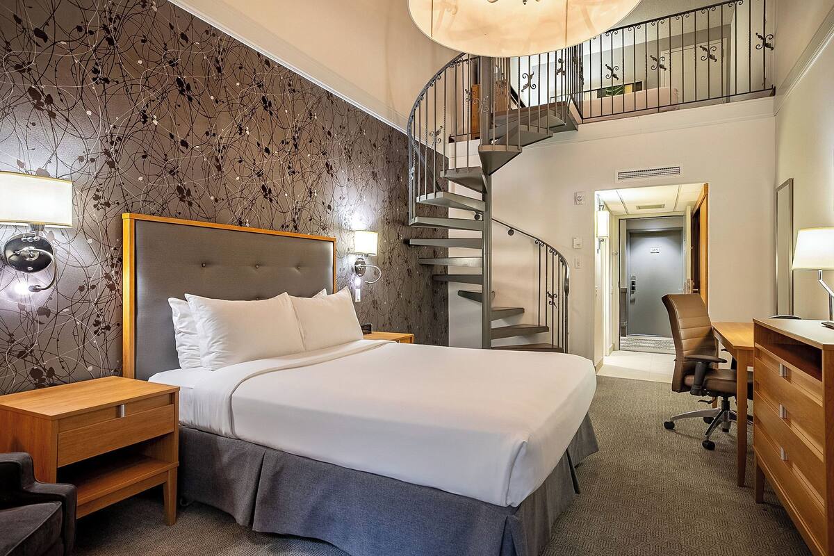 A modern hotel room with a large bed, a desk, a chair, a nightstand, a staircase, and a view of the hallway.