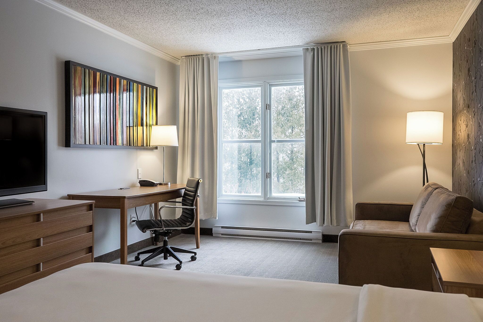 A hotel room with a bed, a desk with a chair, a TV, a window with curtains, and a lamp.