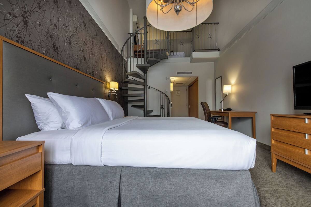 A modern hotel room with a large bed, a wooden nightstand, a desk with a chair, a TV, and a spiral staircase leading to an upper level.