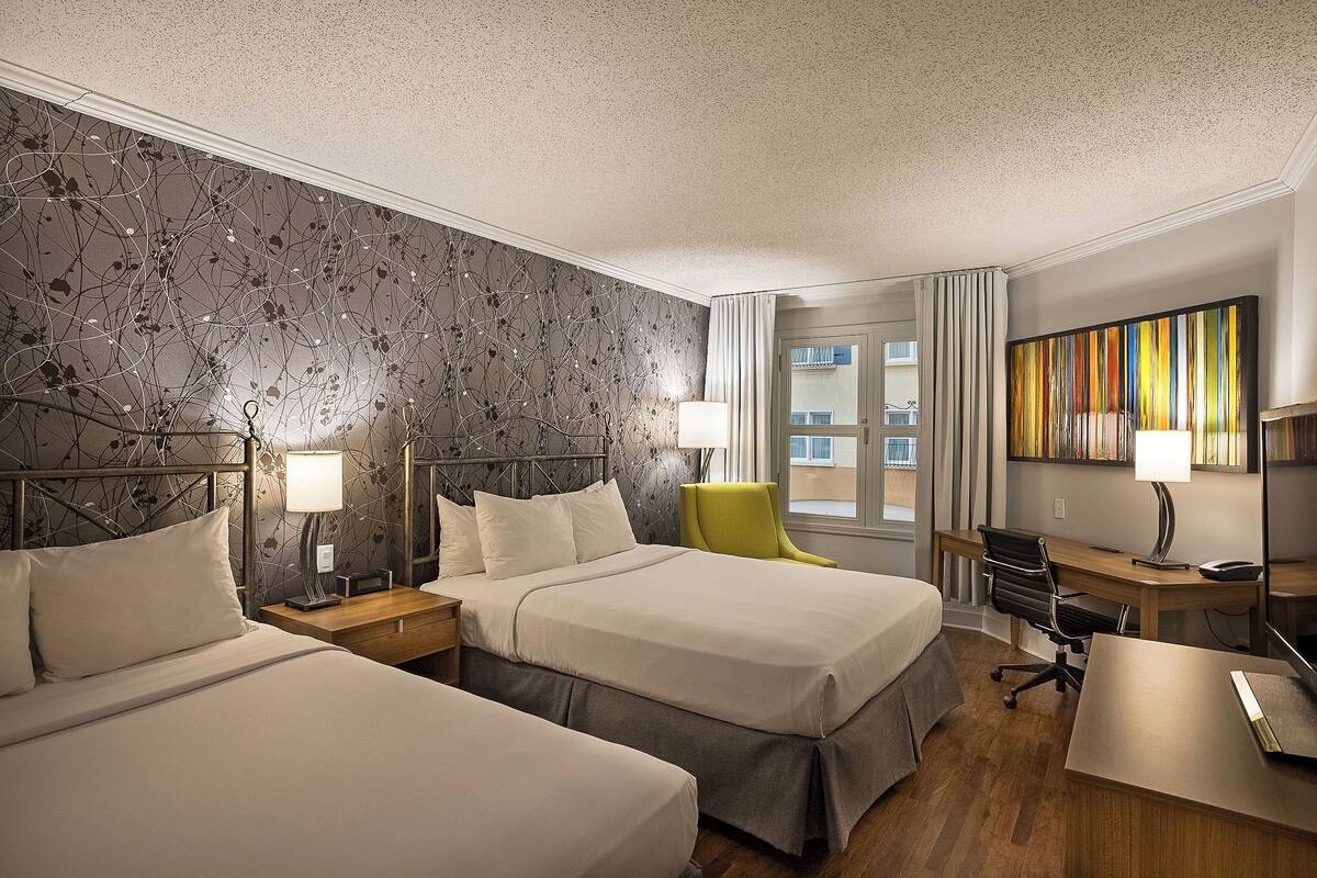 A hotel room with two beds, a desk, a chair, and a large window with curtains.