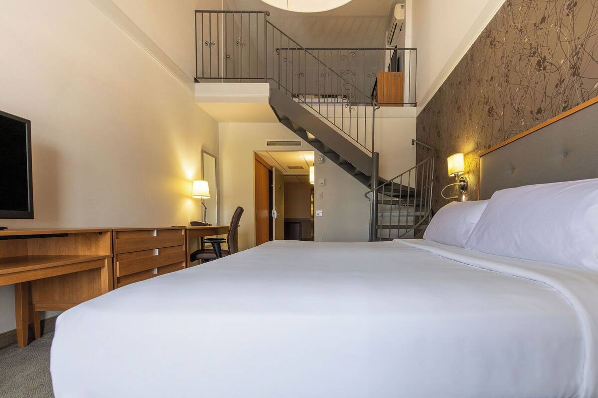 A hotel room with a large bed, wooden desk, chair, and a staircase leading to an upper level.