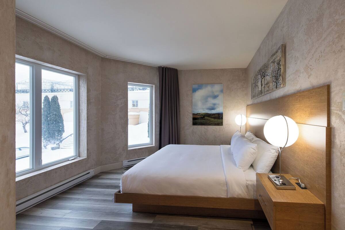 A modern hotel room with a large bed, bedside table, and a view of the outside through large windows.