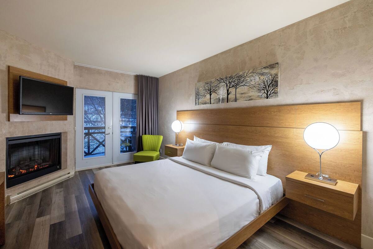 A hotel room with a large bed, a fireplace, a TV mounted on the wall, a nightstand with a lamp, and a window with a view of trees.