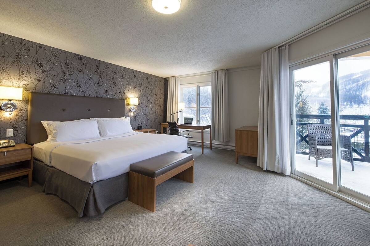 A hotel room with a large bed, a desk, a chair, and a balcony with outdoor furniture.