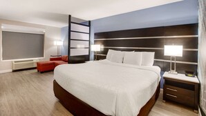 A modern hotel room with a large bed, a red sofa, a desk with a lamp, and a large window.