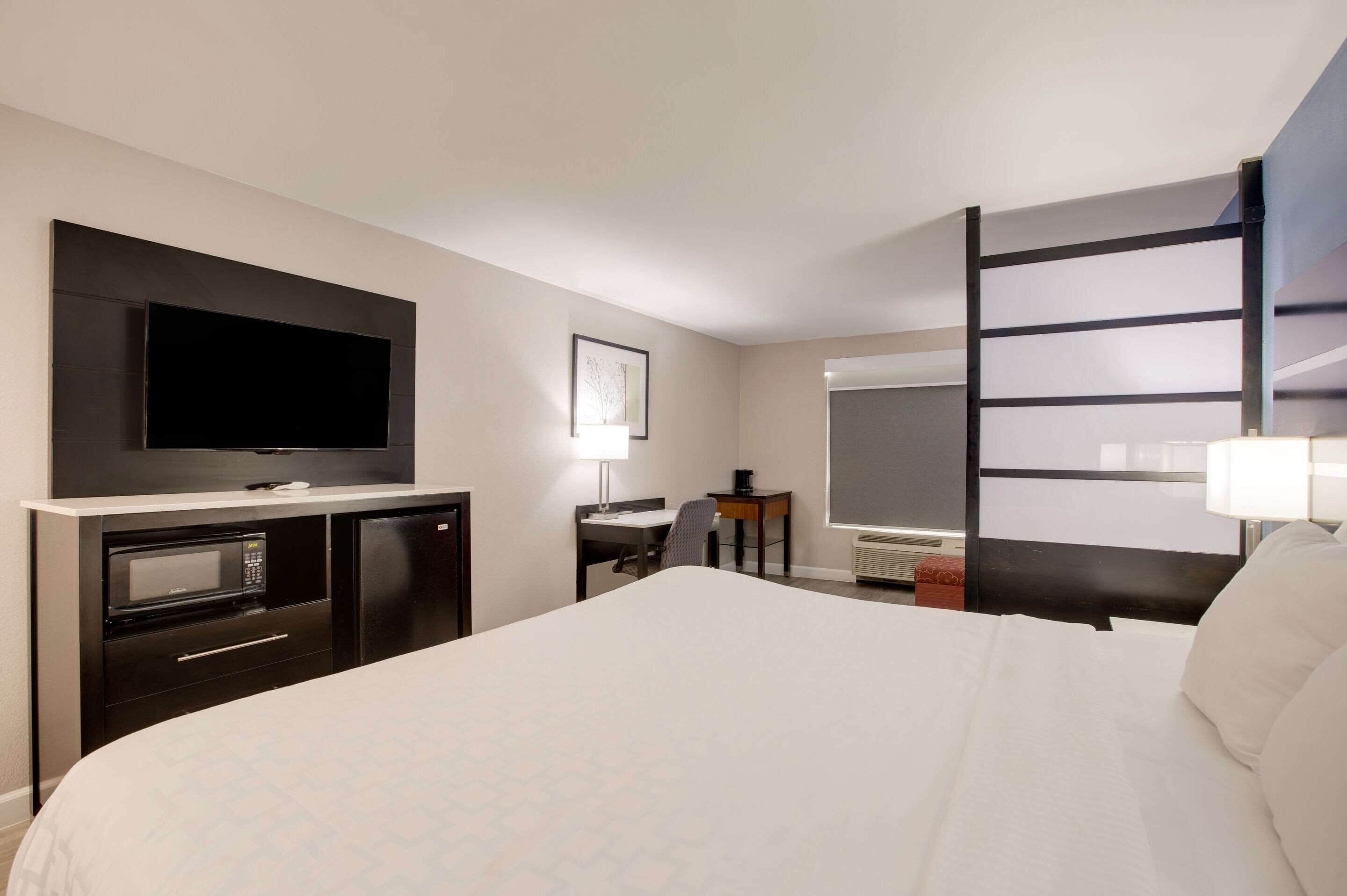 A hotel room with a large bed, a flat-screen TV mounted on the wall, a desk with a chair, a small table with a lamp, and a large mirror.