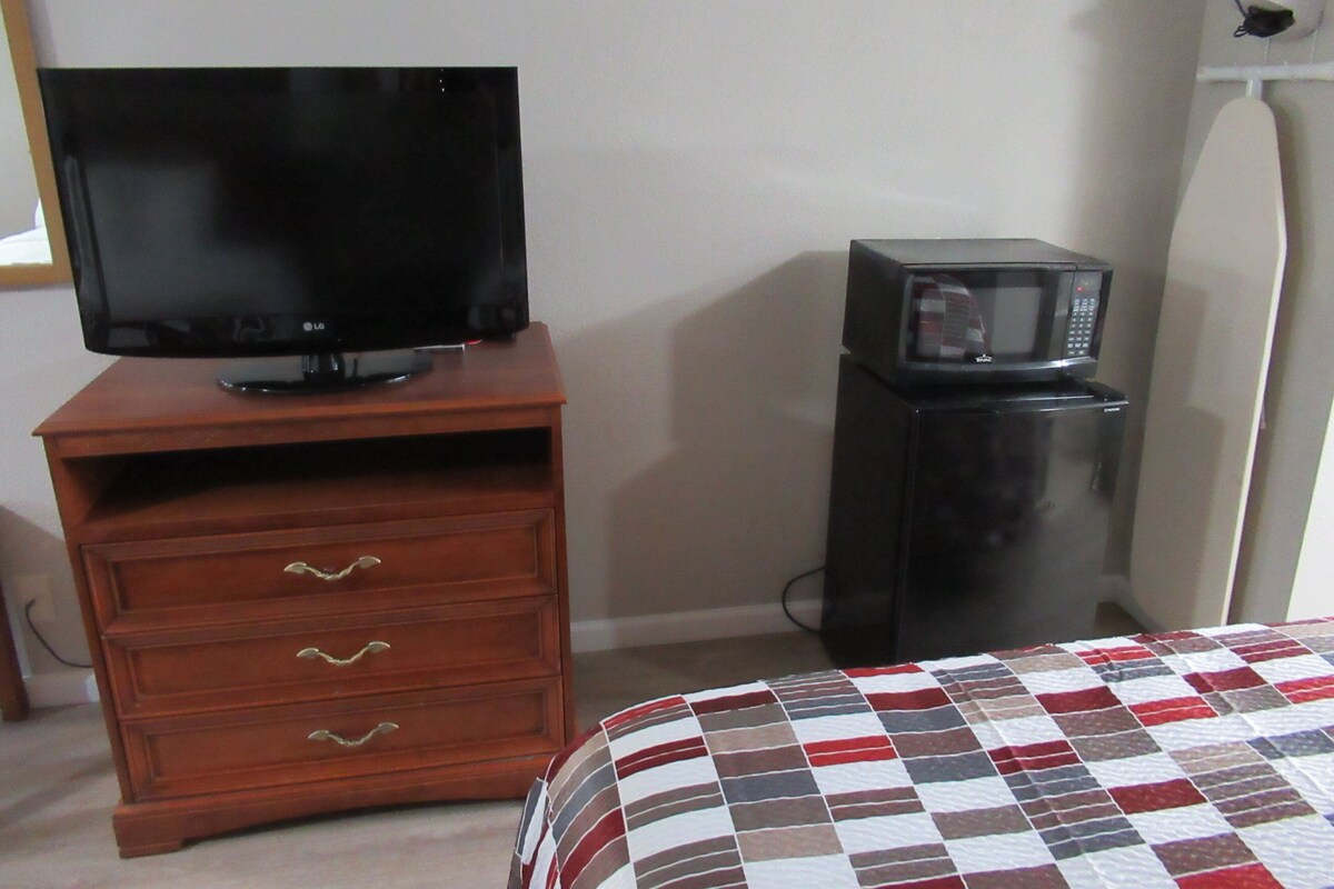 A room with a wooden dresser, a television, a microwave, and a bed with a checkered bedspread.