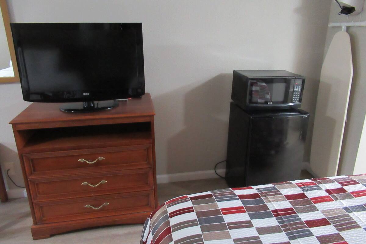 Ein Zimmer mit einem Holzkasten, einem Fernseher, einer Mikrowelle und einem Bett mit karierter Bettdecke.