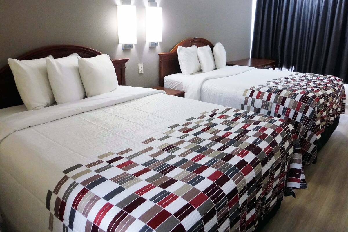 Ein Hotelzimmer mit zwei Betten, jeweils mit weißer Bettwäsche und einer karierten Decke. Über den Betten befinden sich Wandleuchten und am Fenster ein dunkler Vorhang.