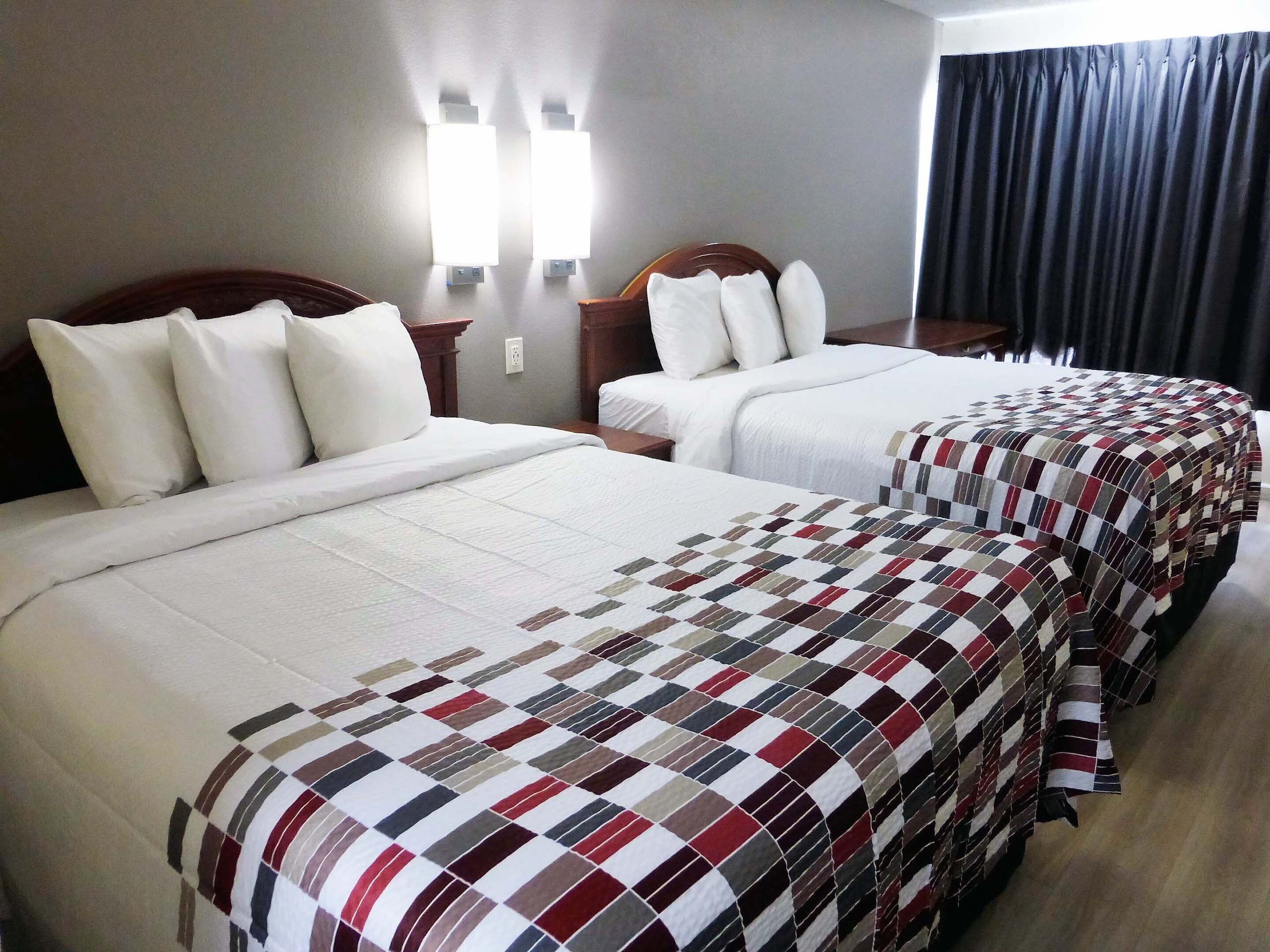A hotel room with two beds, each with white bedding and a checkered blanket. There are wall-mounted lights above the beds and a dark curtain on the window.