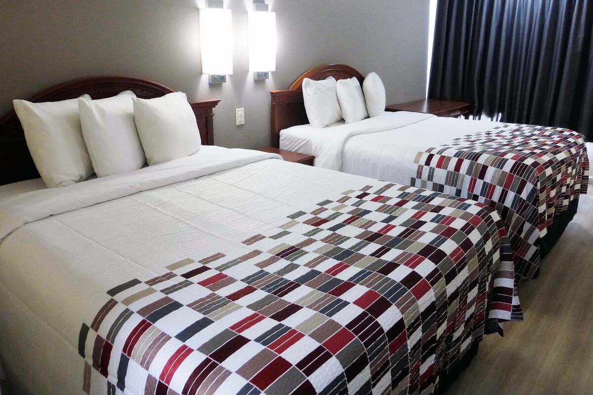 A hotel room with two beds, each with white bedding and a checkered blanket. There are wall-mounted lights above the beds and a dark curtain on the window.