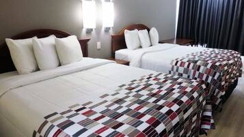 A hotel room with two beds, each with white bedding and a checkered blanket. There are wall-mounted lights above the beds and a dark curtain on the window.