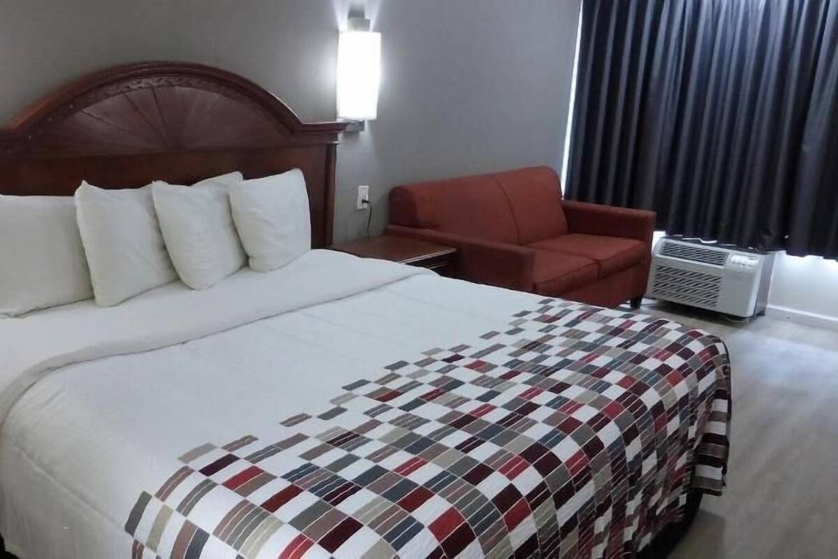 Ein Hotelzimmer mit einem großen Bett, einem Holzkopfende, einem roten Sessel, einem Nachttisch und einer Wand-Klimaanlage.