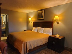 A hotel room with a large bed, two bedside lamps, a nightstand with a phone, and a painting on the wall. - Garden Inn & Extended Stay Shepherdsville / Louisville (Shepherdsville)
