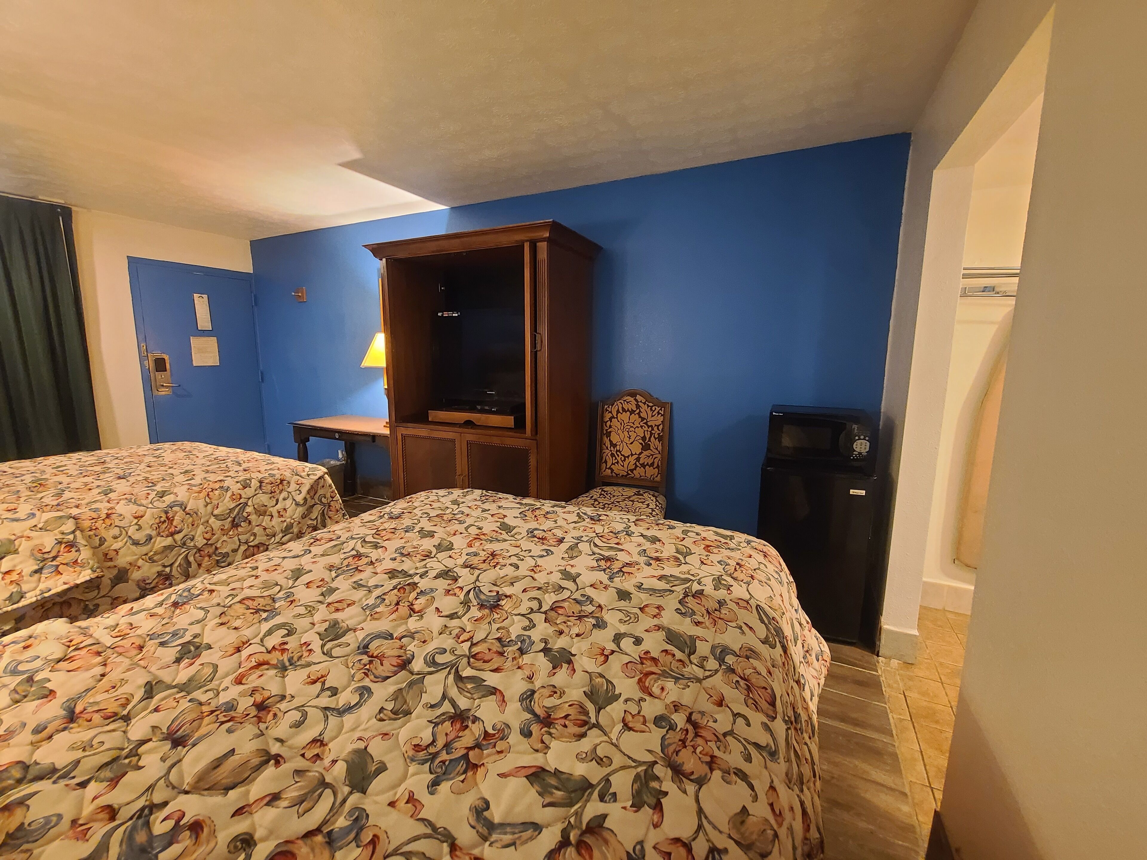A hotel room with two beds, a wooden cabinet, a desk with a lamp, and a refrigerator.