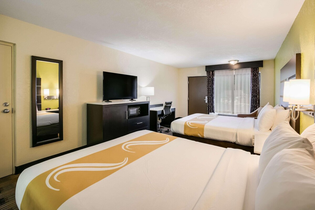 A hotel room with a large bed, a television, a desk with a chair, and a window with curtains.