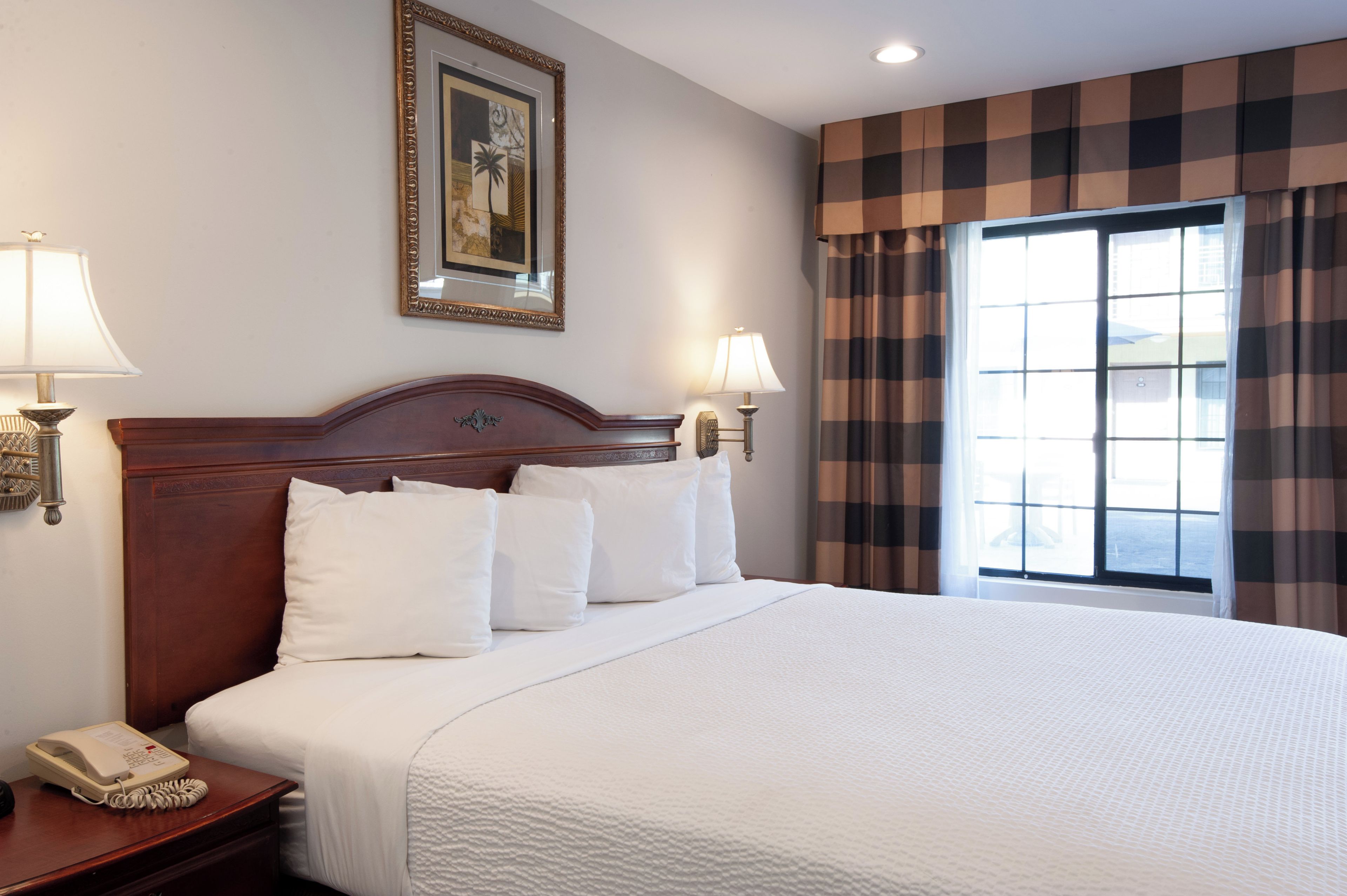 A hotel room with a large bed, two bedside lamps, a wooden headboard, a framed artwork on the wall, and a window with checkered curtains.