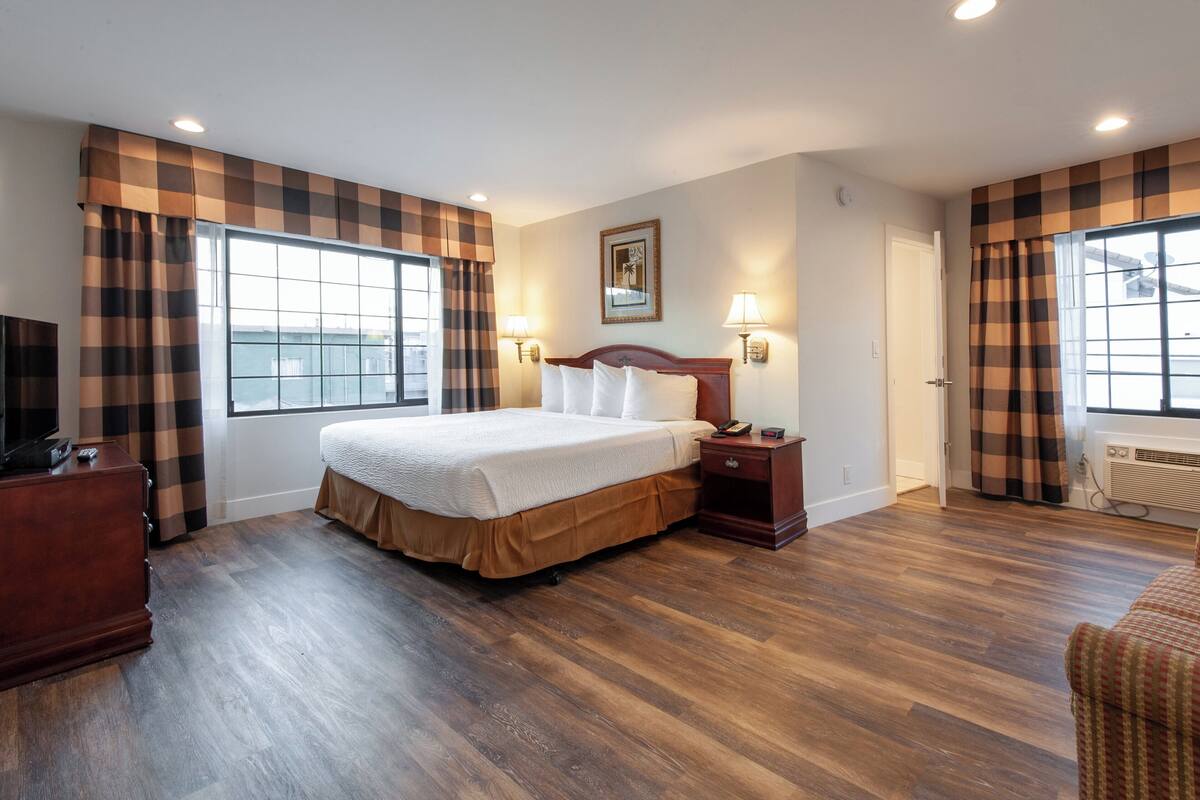 Una habitación de hotel con una cama grande, dos lámparas de noche, un aparador de madera y una ventana con cortinas a cuadros.
