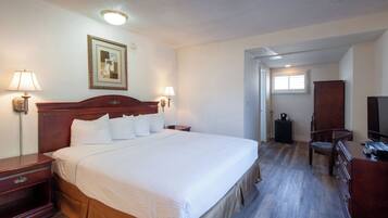 A hotel room with a large bed, bedside lamps, a wooden headboard, a desk with a chair, a dresser, a television, and a framed picture on the wall.
