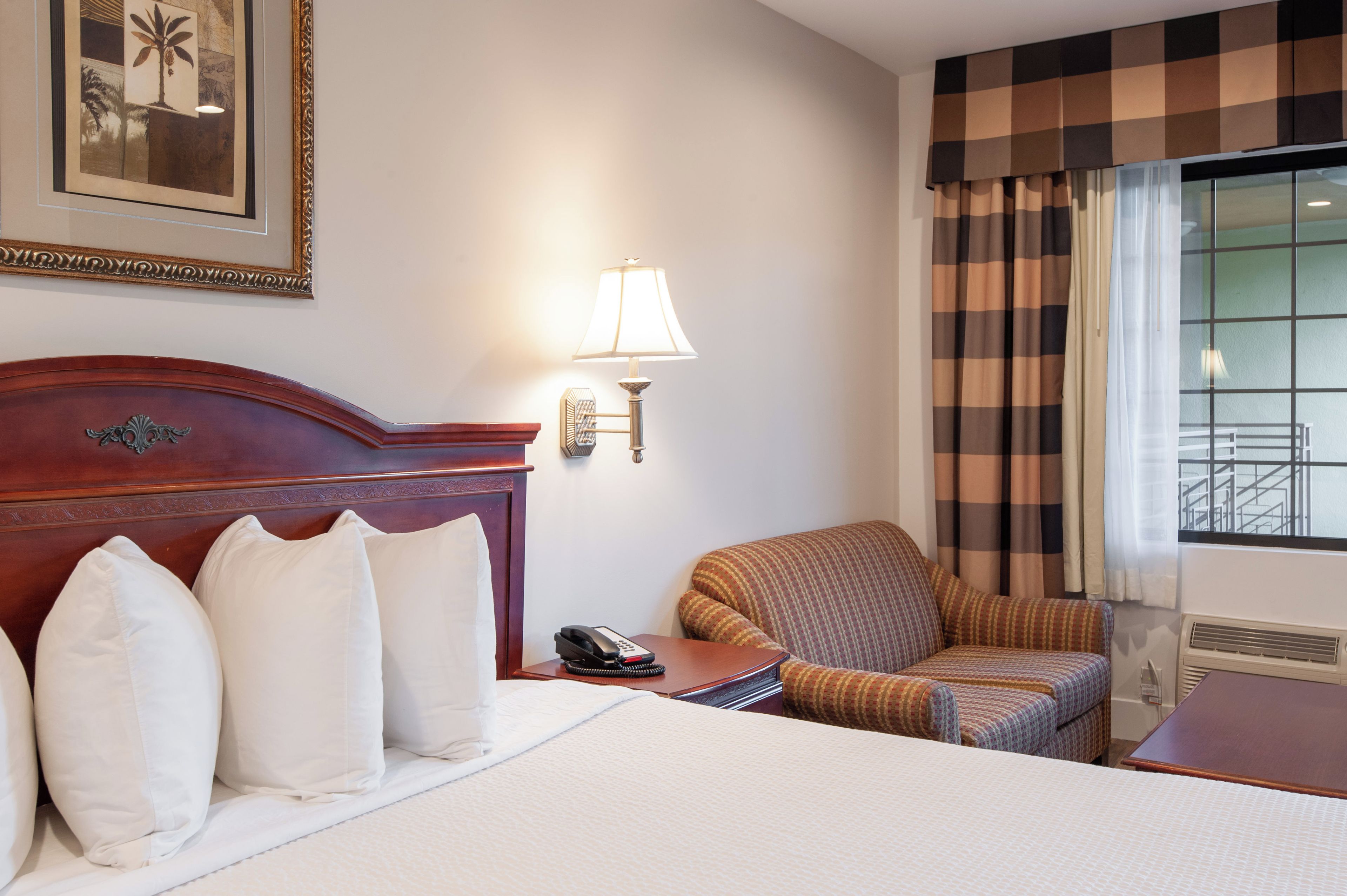 Una habitación de hotel con una cama, una silla, una mesita de noche con lámpara y un cuadro en la pared.