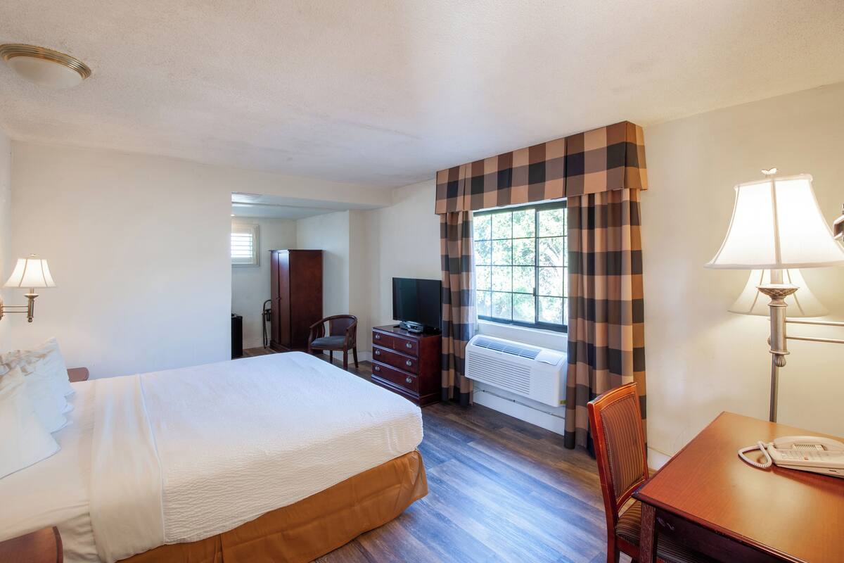 Una habitación de hotel con cama, escritorio con teléfono, silla, cómoda y ventana con cortinas, además de un equipo de aire acondicionado.