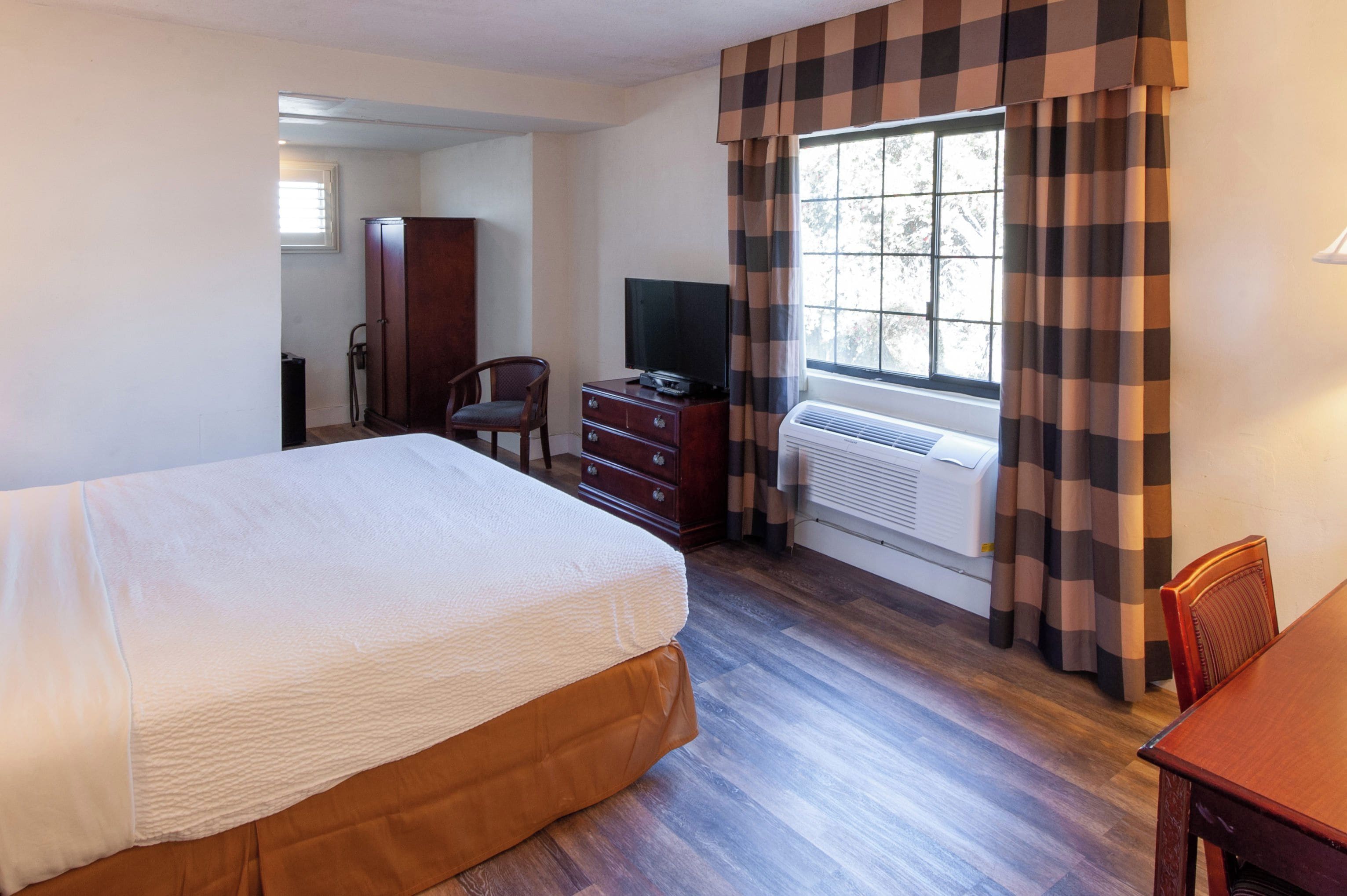 Una habitación de hotel con una cama grande, un aparador de madera, un televisor y una ventana con cortinas a cuadros.