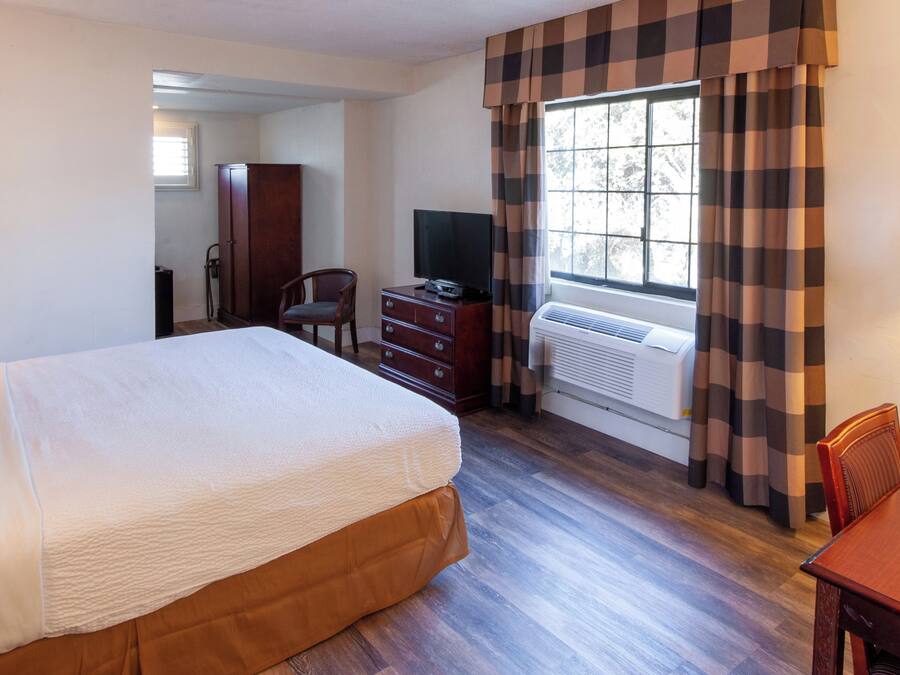 Quarto de hotel com uma cama grande, cômoda de madeira, televisão e janela com cortinas xadrez.