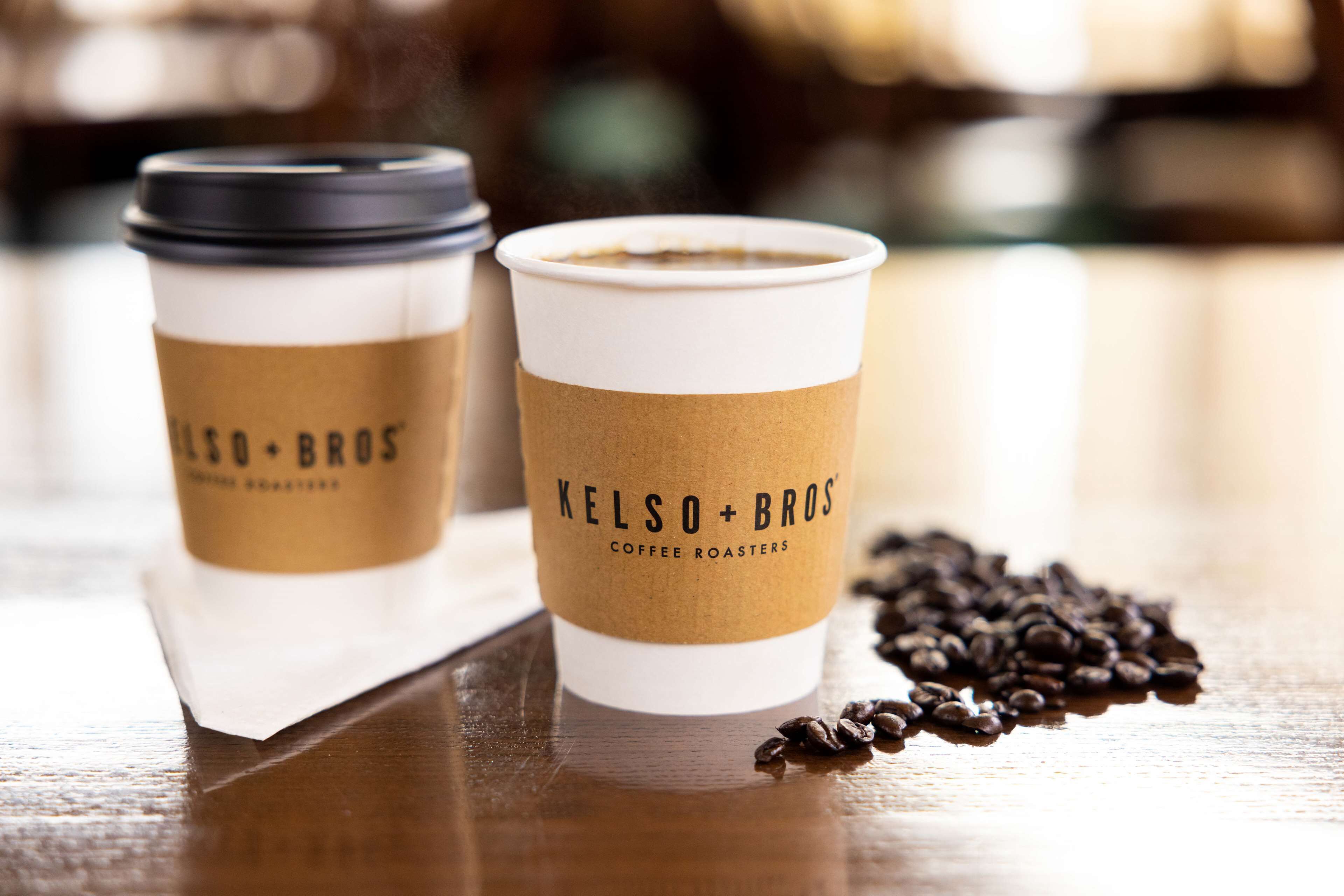 Två kaffekoppar med logotypen för ”Kelso+Bros Coffee Roasters” placerade på en träyta, omgiven av utspridda kaffebönor.