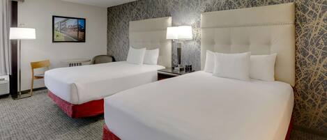 A hotel room with two beds, a small table, a chair, a lamp, and a painting on the wall.