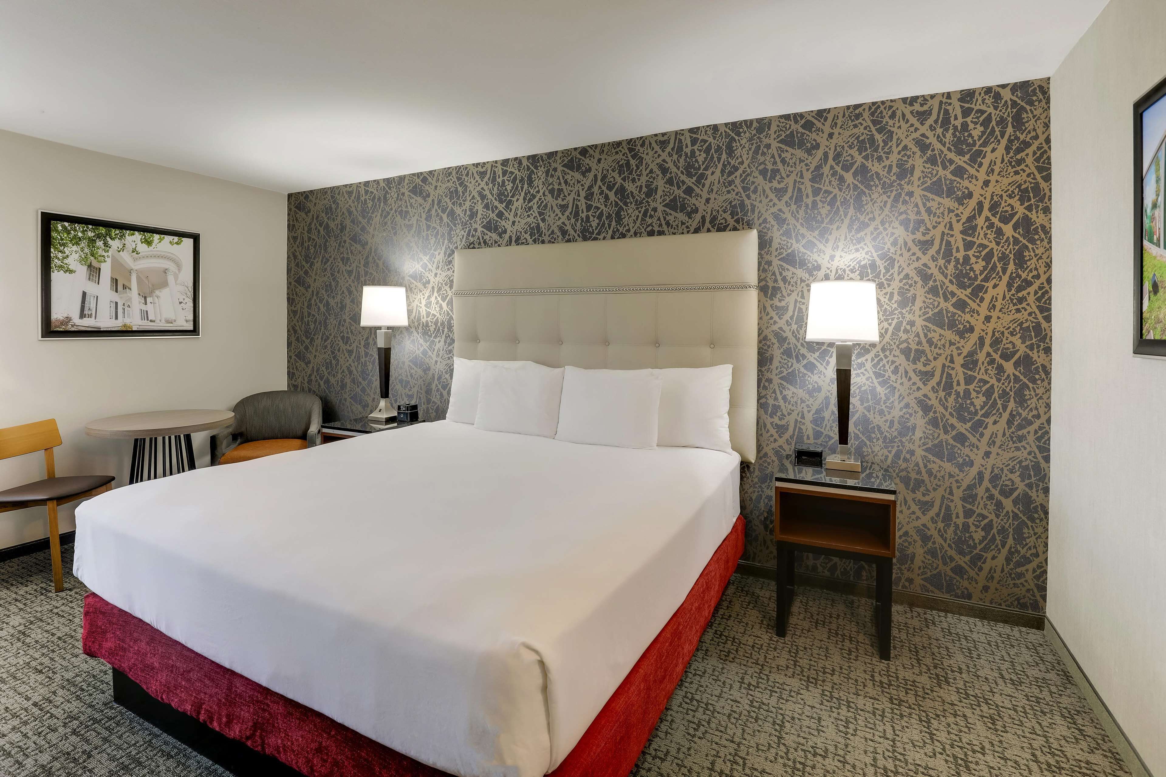Habitación de hotel con una cama grande, dos mesitas de noche con lámparas, una silla, una pequeña mesa y un cuadro en la pared.