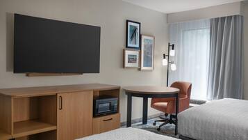 Habitación de hotel con televisión de pantalla plana, escritorio con microondas, una silla, una mesita y dos camas.
