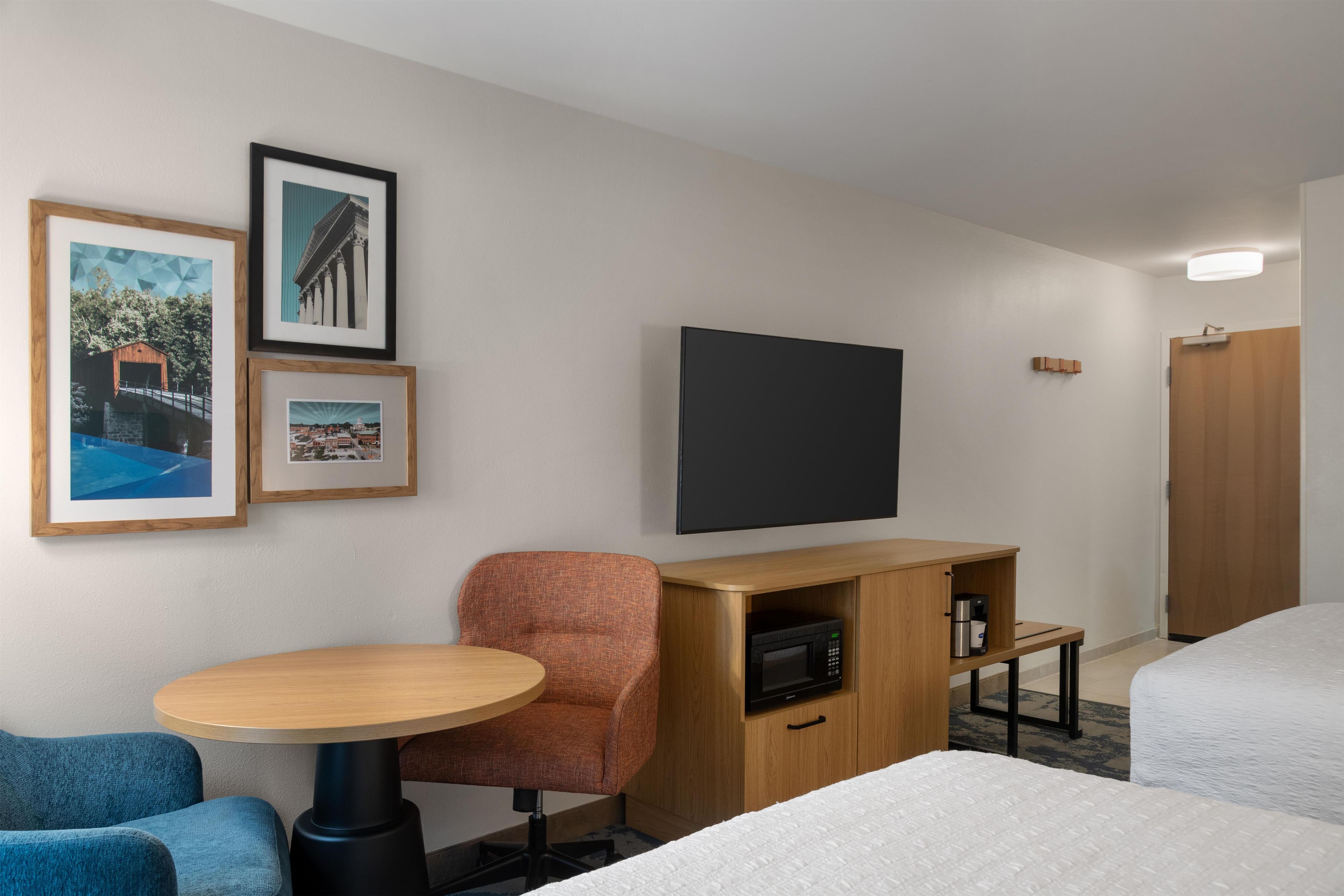 A hotel room with a bed, a desk with a microwave, a chair, a small table, and framed pictures on the wall.