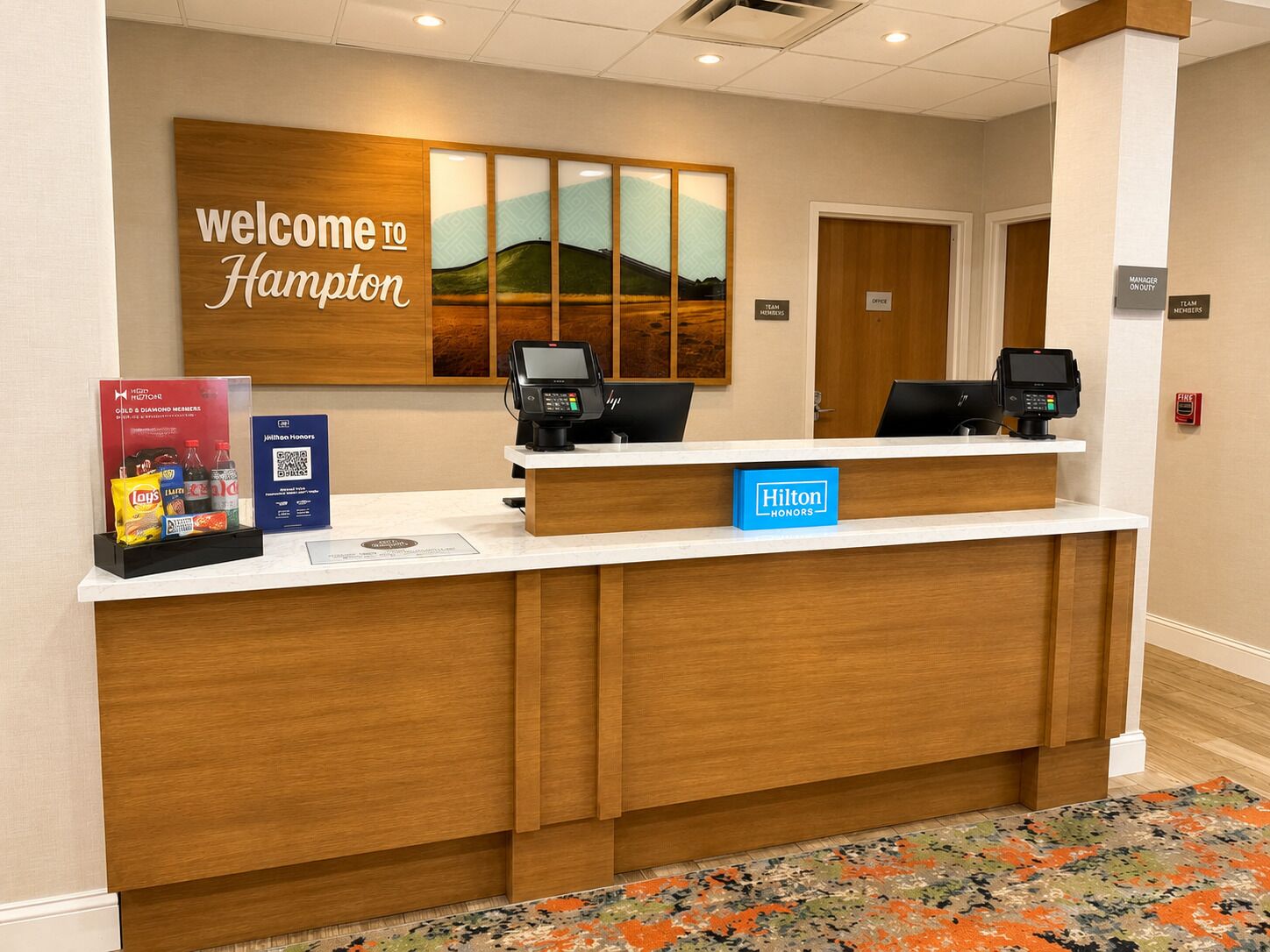 Mostrador de recepción de hotel con un cartel que dice 'Welcome to Hampton', dos monitores de computadora, una tarjeta 'Hilton Honors' y un cartel que dice 'Team Members'.