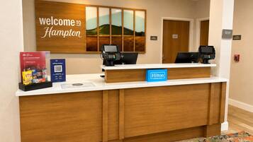 Mostrador de recepción de hotel con un cartel que dice 'Welcome to Hampton', dos monitores de computadora, una tarjeta 'Hilton Honors' y un cartel que dice 'Team Members'.