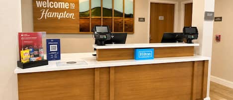 Mostrador de recepción de hotel con un cartel que dice 'Welcome to Hampton', dos monitores de computadora, una tarjeta 'Hilton Honors' y un cartel que dice 'Team Members'.