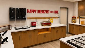 A breakfast area with a sign that reads 'HAPPY BREAKFAST STARTS HERE.' There are black cabinets with utensils, a red oatmeal dispenser, and a fruit basket on the counter.
