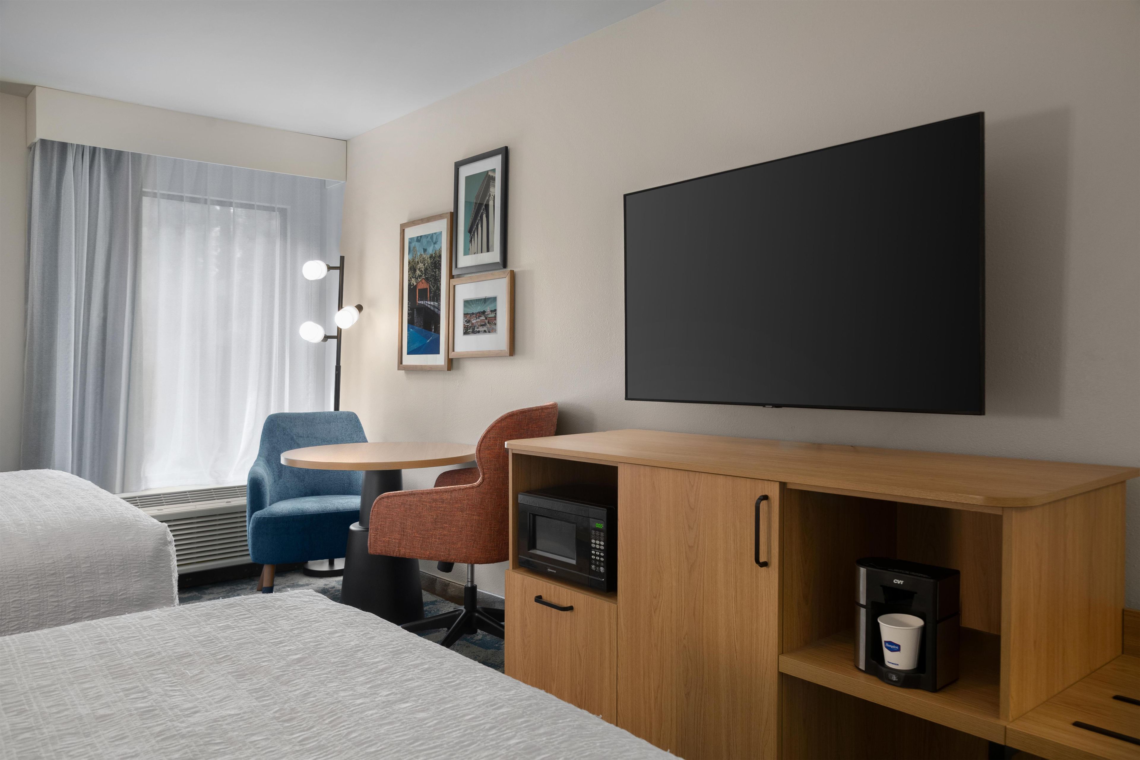 A hotel room with a bed, a desk with a microwave, a coffee machine, a TV, and a chair.