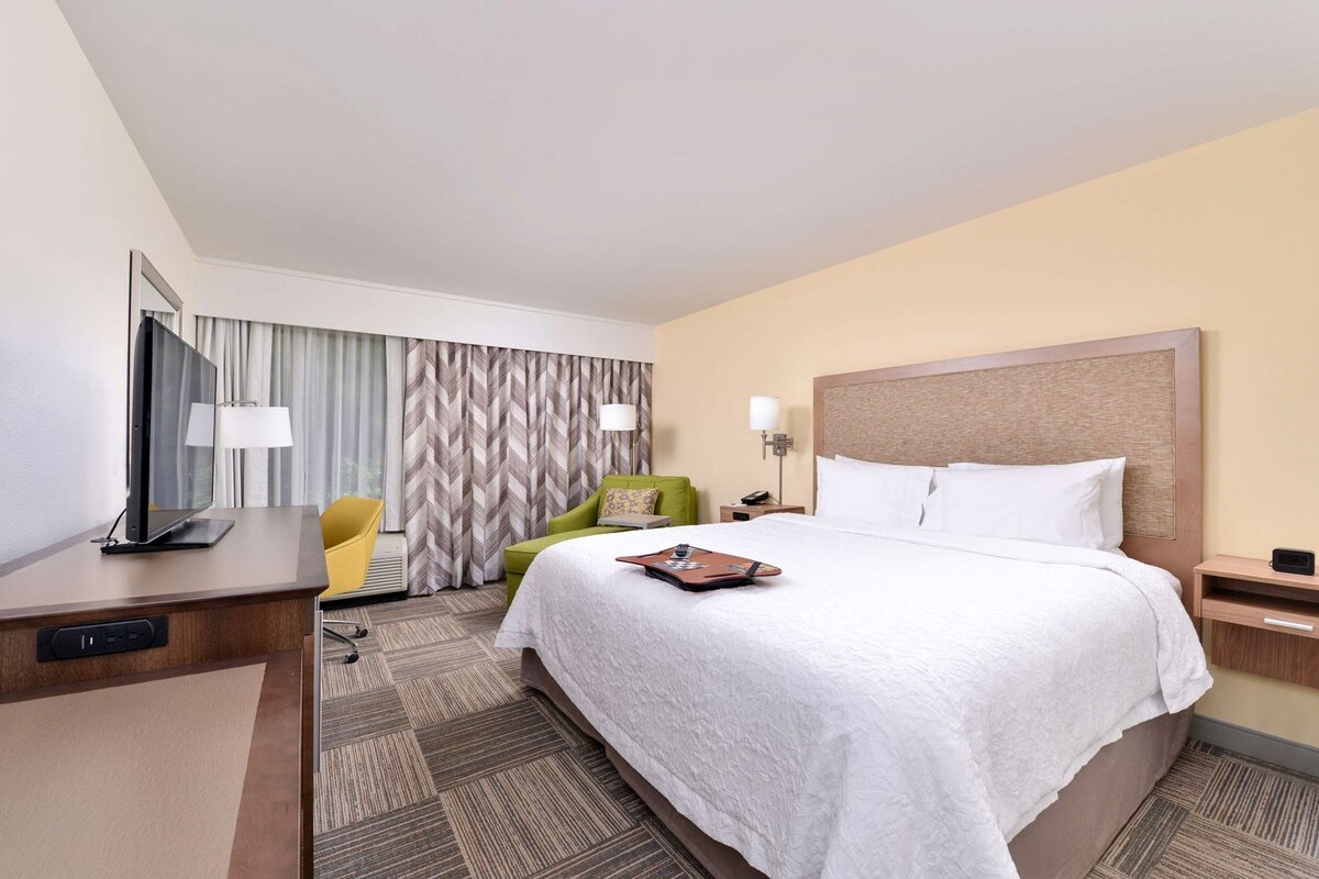 A hotel room with a bed, a desk with a computer, a chair, a lamp, and a television.
