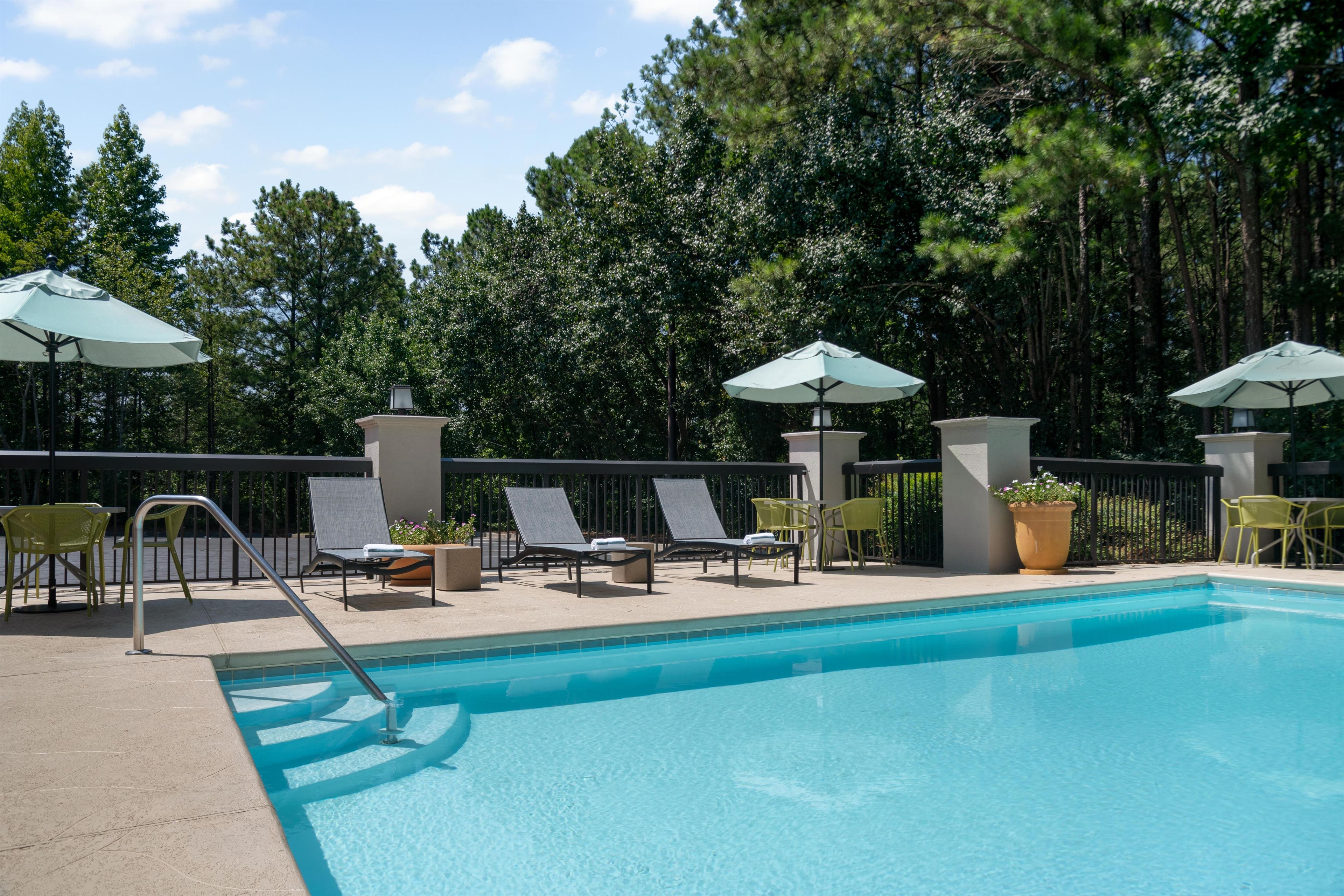 A swimming pool with lounge chairs and umbrellas, surrounded by a fence and trees.