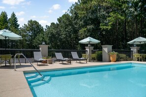 Une piscine entourée d’une clôture et d’arbres, avec des chaises longues et des parasols à proximité.
