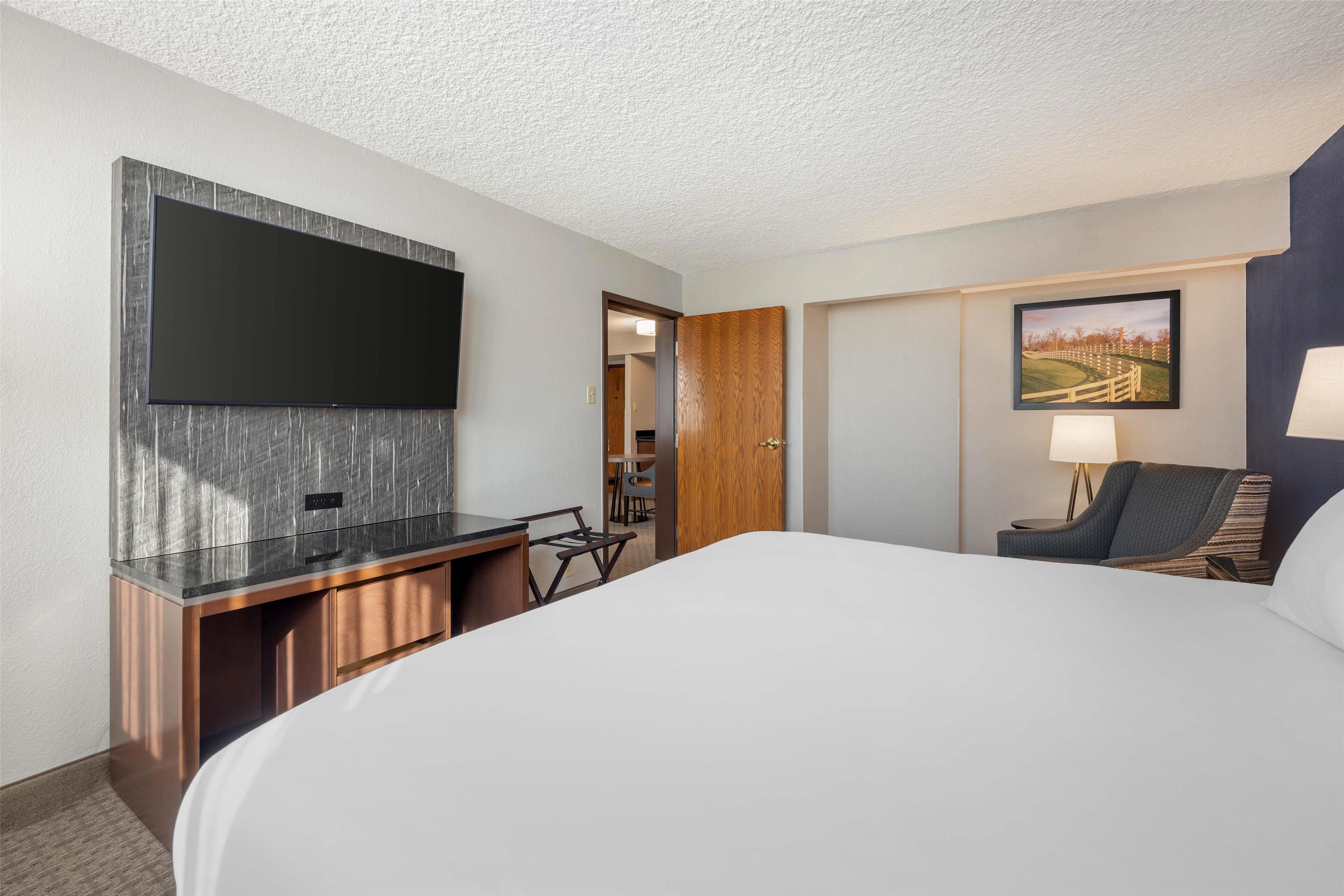 A hotel room with a large bed, a flat-screen TV mounted on the wall, a desk with a chair, a lamp, and a painting on the wall.